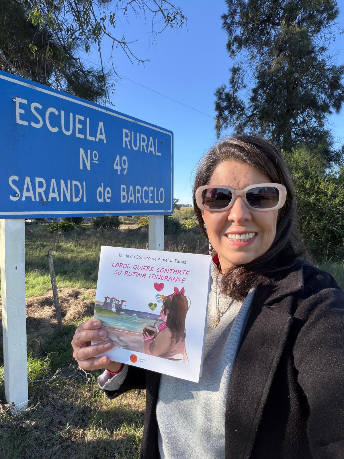 Maria do Socorro com o livro Carol ao lado da placa da Escuela Rural Nº 49 Sarandi de Barcelo